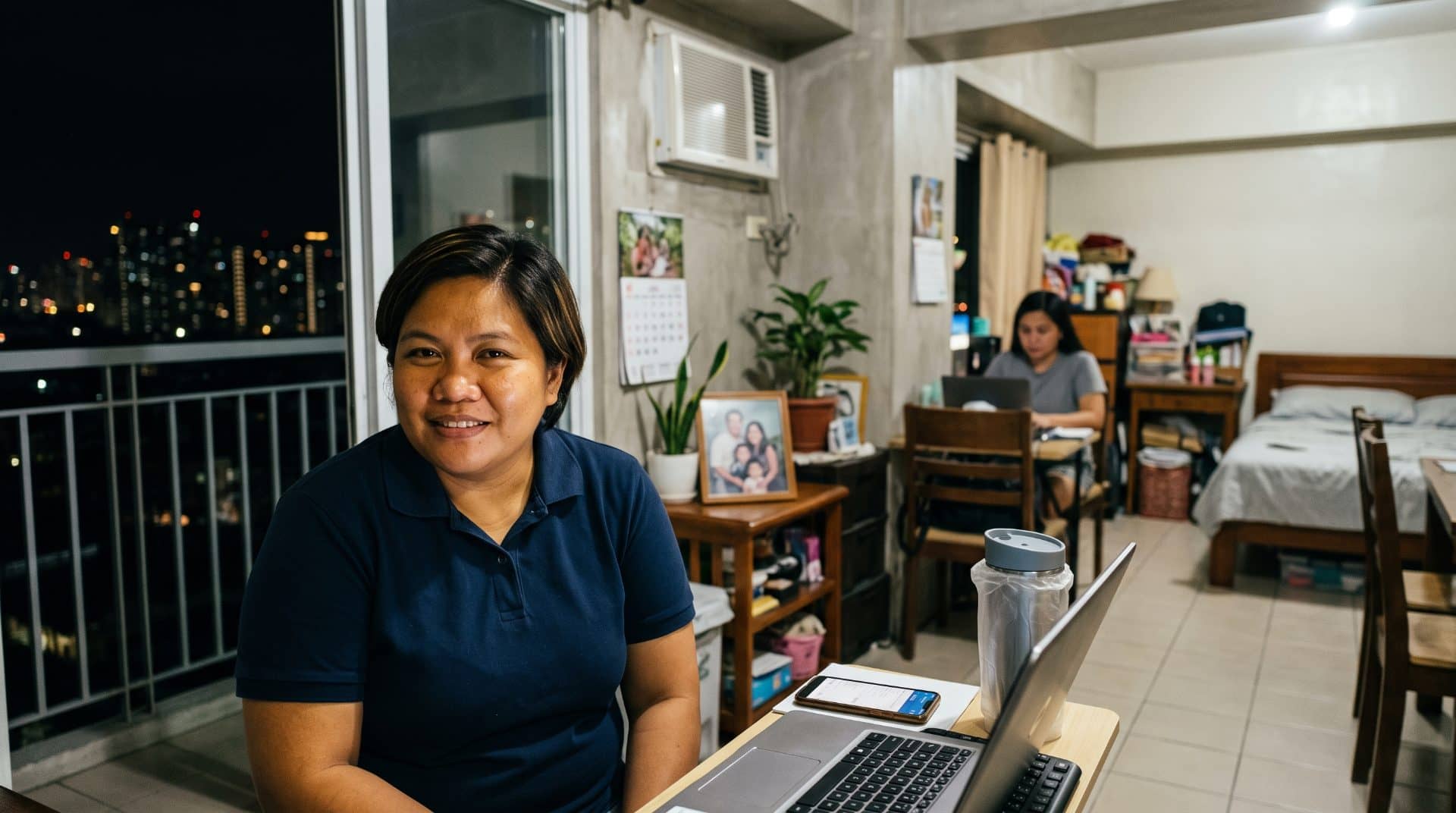 Two women review work at a desk by a window at night, a phone and keyboard nearby.