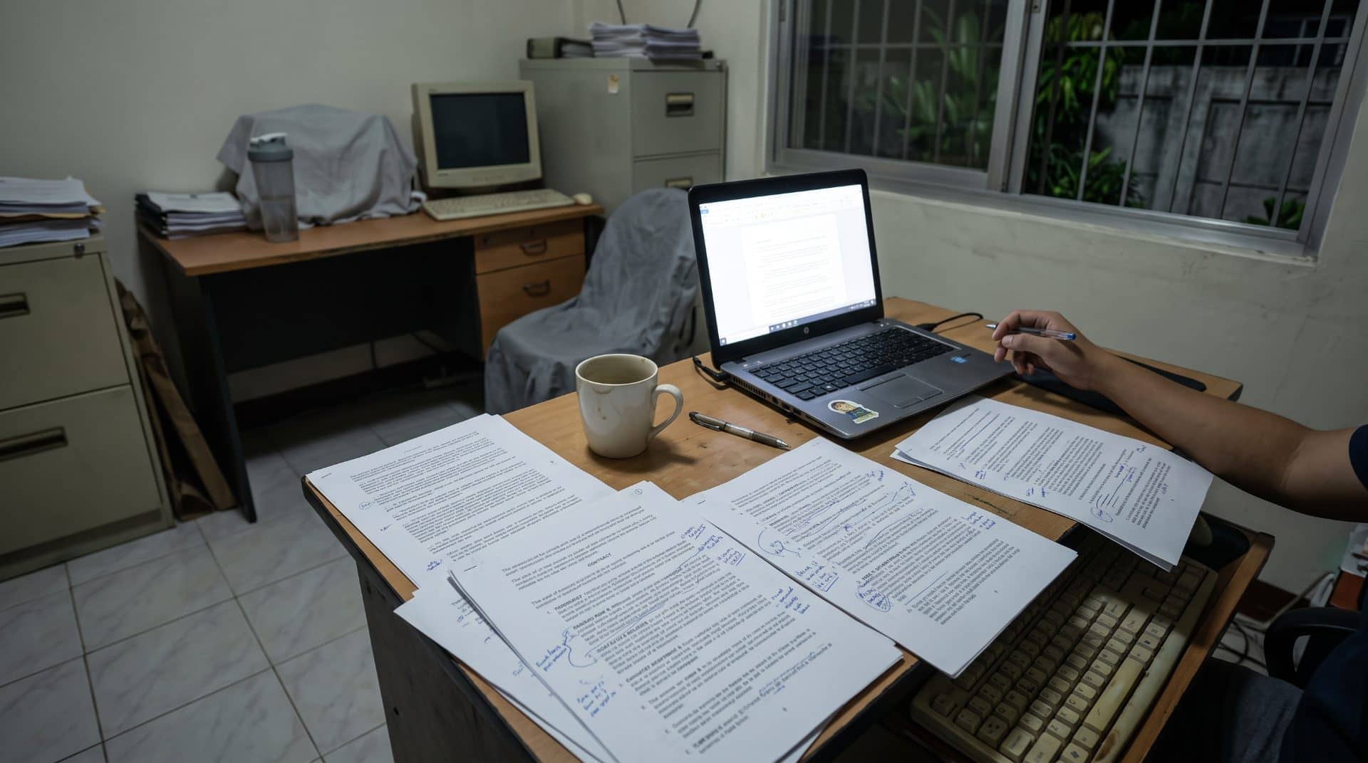 Marked-up contract pages and cold coffee on a desk at night, lit from above.